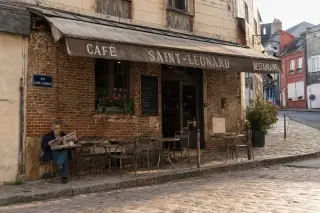 Quiet morning outside an old café in Saint-Léonard, Honfleur, a local reading at a table with warm golden sunlight on cobblestones and aged façade