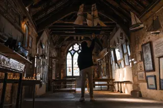 A person stands inside a historic chapel at dawn, gently touching a model ship hanging from the ceiling, surrounded by maritime ex-votos and soft golden light.