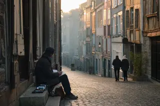 Solitary painter sketching on a cobbled street in Honfleur at dawn, bathed in gentle golden light with historic pastel buildings in the background.