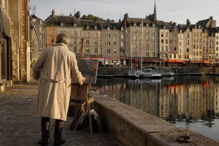 Artist setting up an easel on a quiet historic quay at sunset, colorful old warehouses reflected in the still port water, soft golden spring light.