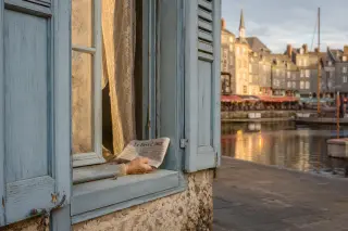 Honfleur old port window with blue shutters, a hand holding a newspaper on the sill, soft golden evening light and blurred colorful facades reflected in the basin