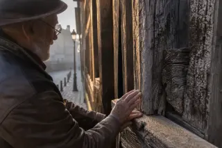 Close-up of elderly hands on Sainte-Catherine church’s weathered wooden beams in soft dawn light, mist and cobblestones in the background.