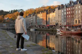 Lone person on a stone jetty overlooking Honfleur's old harbor at sunset, softly illuminated, quietly blending urban and natural landscapes.