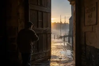 Moment naturaliste dans l’entrée d’un grenier à sel ancien à Honfleur, silhouette locale ouvrant une lourde porte à l’aube, détails de pierres anciennes et lumière dorée sur pavés mouillés